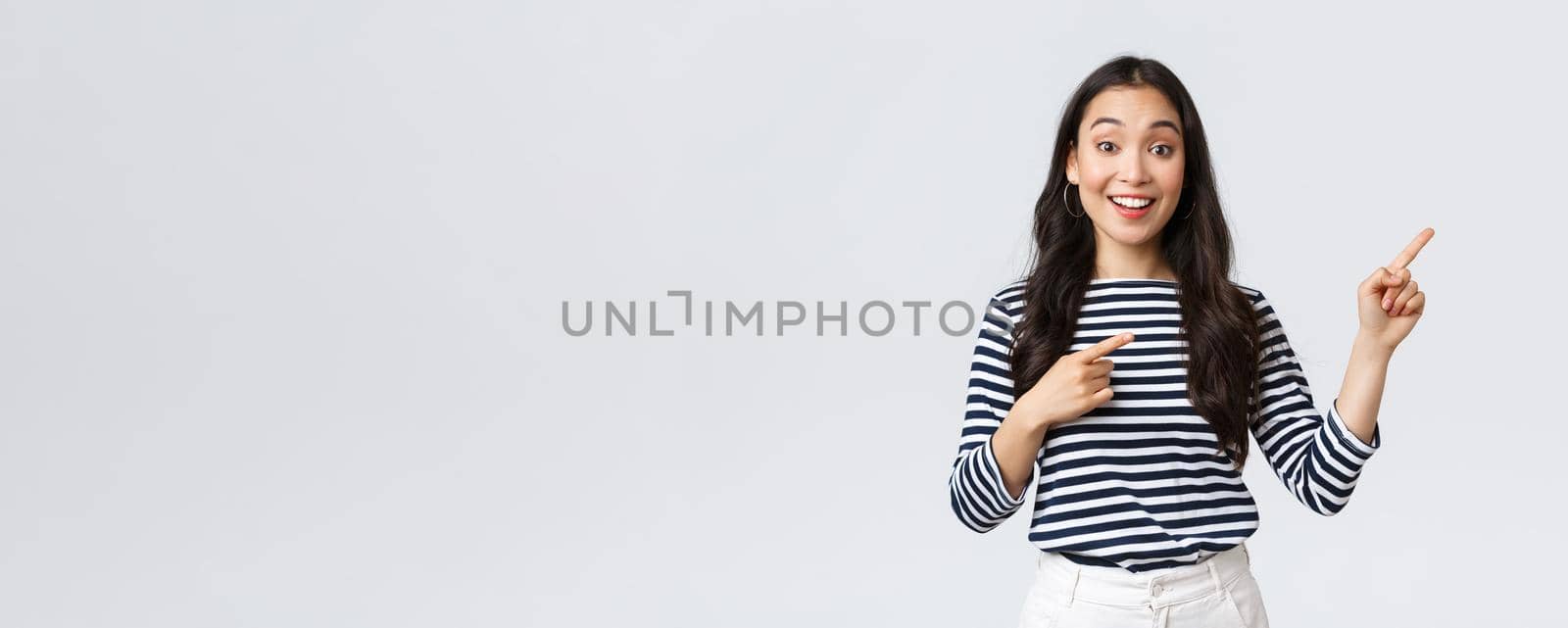 Lifestyle, beauty and fashion, people emotions concept. Surprised happy asian girl standing over white background recommend visit shop, pointing fingers right at banner.