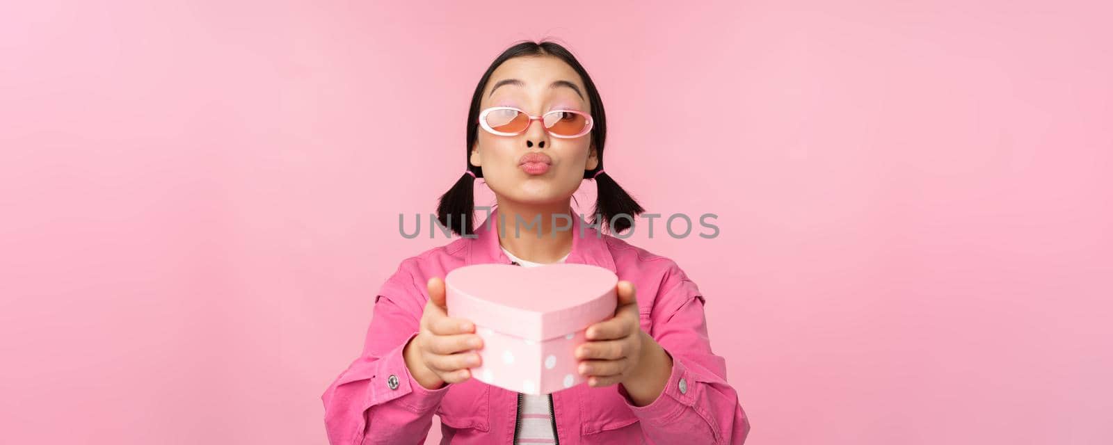 Cute asian girl giving you gift in heart shaped box, kissing and smiling, concept of holiday and celebration, standing over pink background.