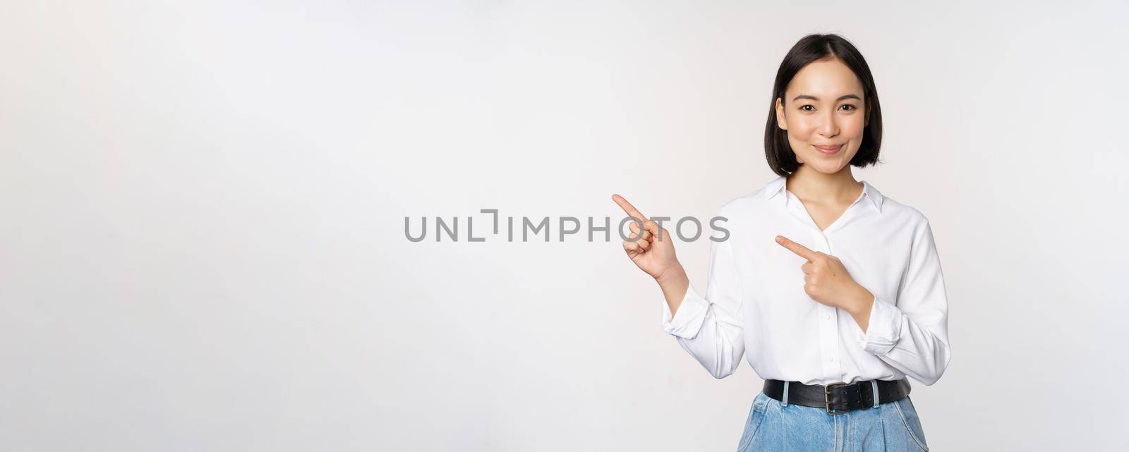 Image of smiling young office lady, asian business entrepreneur pointing fingers left, showing client info, chart of banner aside on copy space, white background.