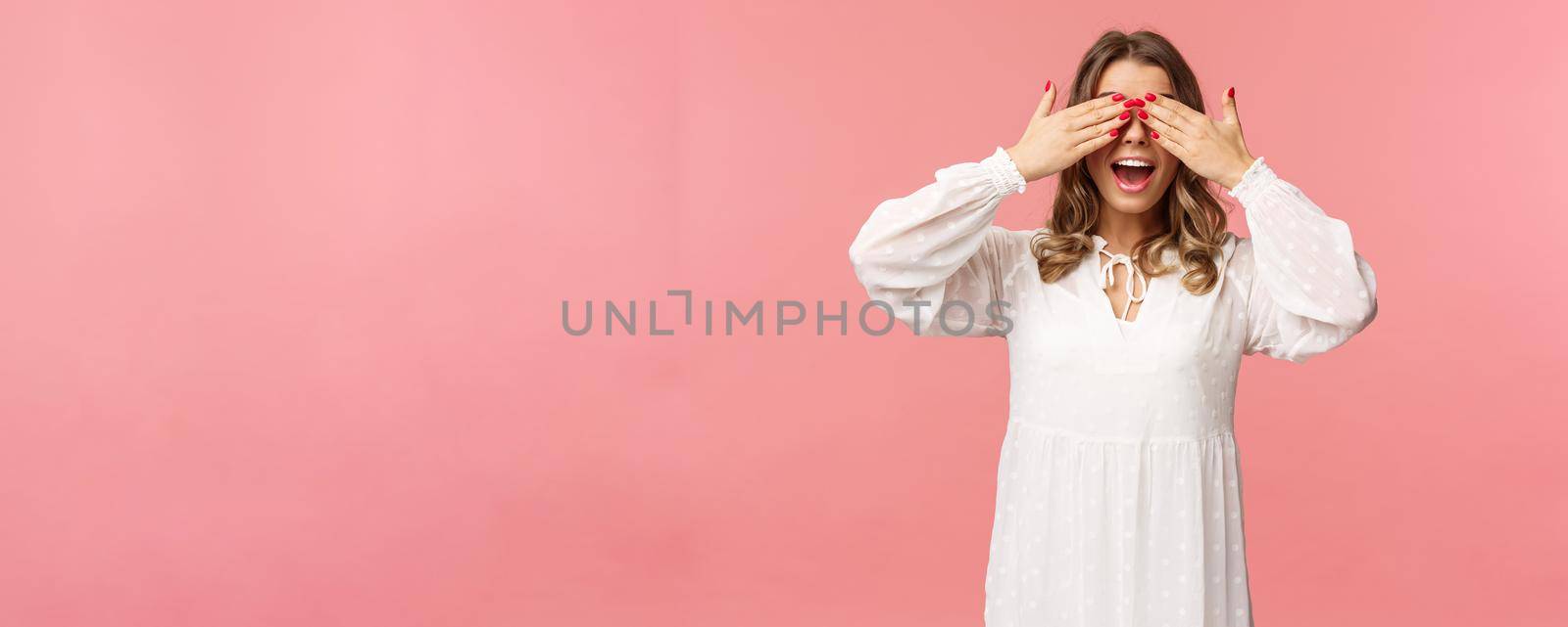 Excited cute blond girl in white spring dress waiting for gifts at surprise party, cover eyes blindfolded standing pink background, open mouth in amazement and anticipation.