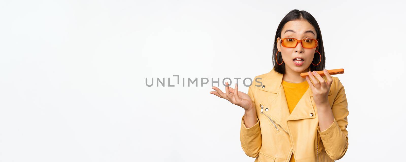 Image of happy asian girl talking on speakerphone, recording, translating her voice with mobile phone app, talking in smartphone dynamic, standing over white background.