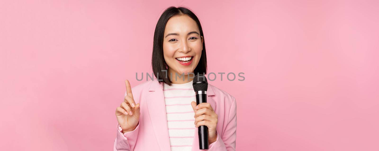 Image of enthusiastic asian businesswoman giving speech, talking with microphone, holding mic, standing in suit against pink studio background by Benzoix