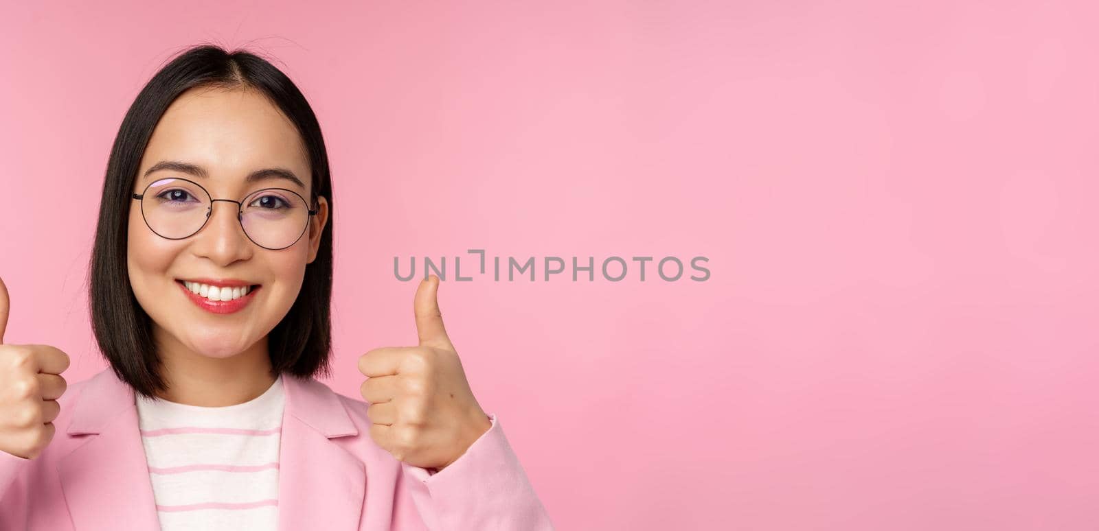 Awesome, congrats. Face of excited asian businesswoman in glasses, smiling pleased, showing thumbs up in approval, standing over pink background by Benzoix