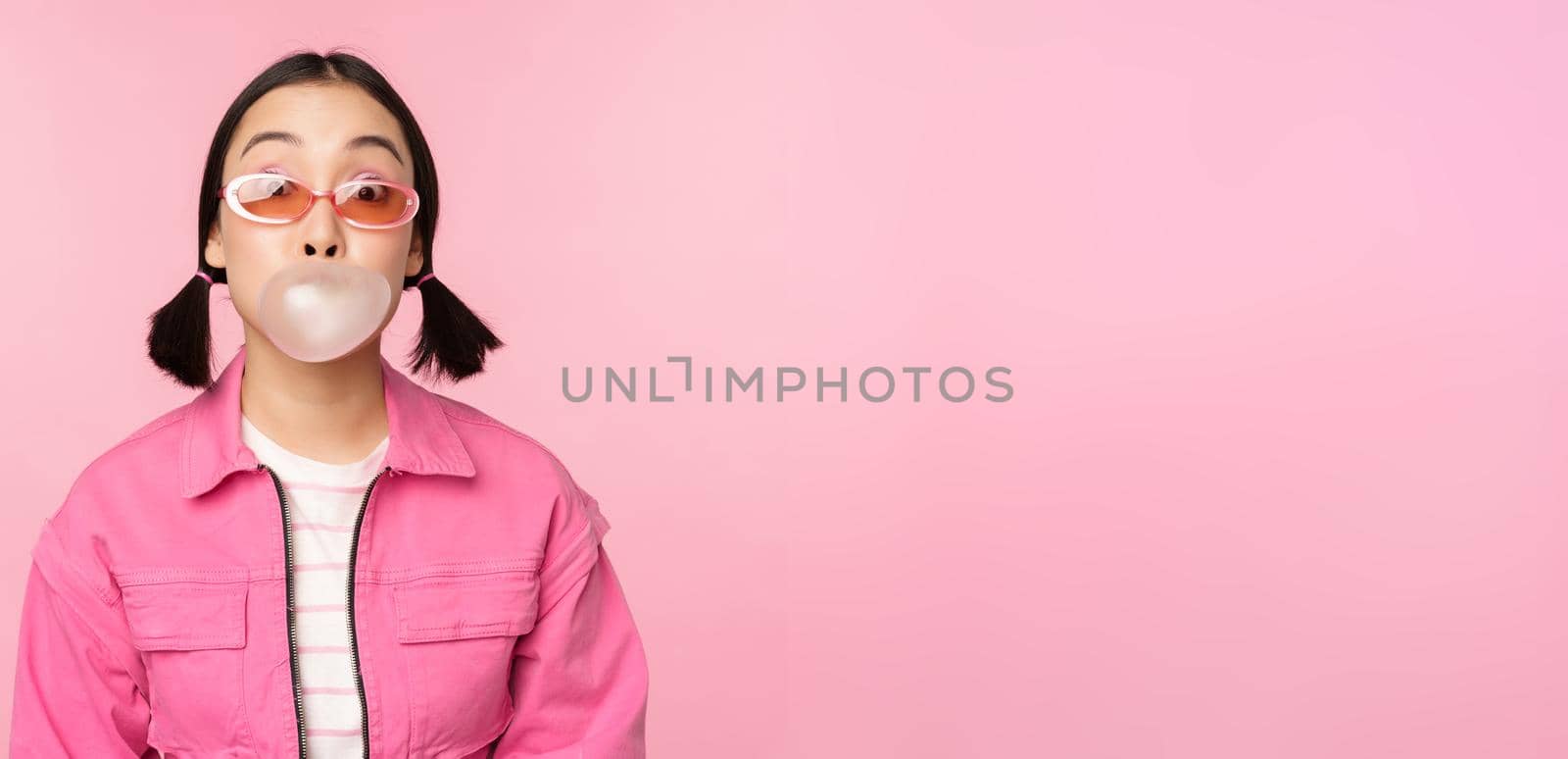 Stylish asian girl blowing bubblegum bubble, chewing gum, wearing sunglasses, posing against pink background by Benzoix