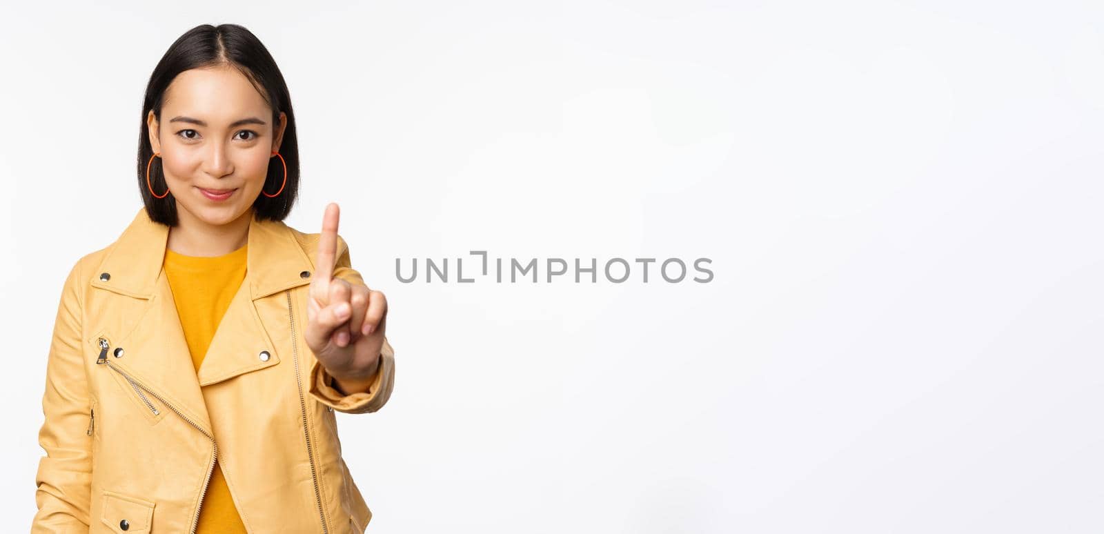 Rule number one. Confident smiling asian woman showing index finger, stop, prohibit gesture, disapprove smth, standing over white background by Benzoix