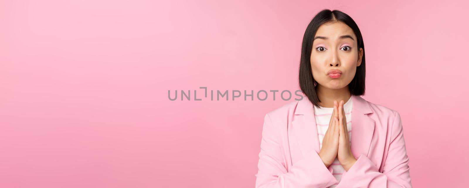 Portrait of asian businesswoman asking for help, say please, standing in praying, begging pose, pink studio background by Benzoix