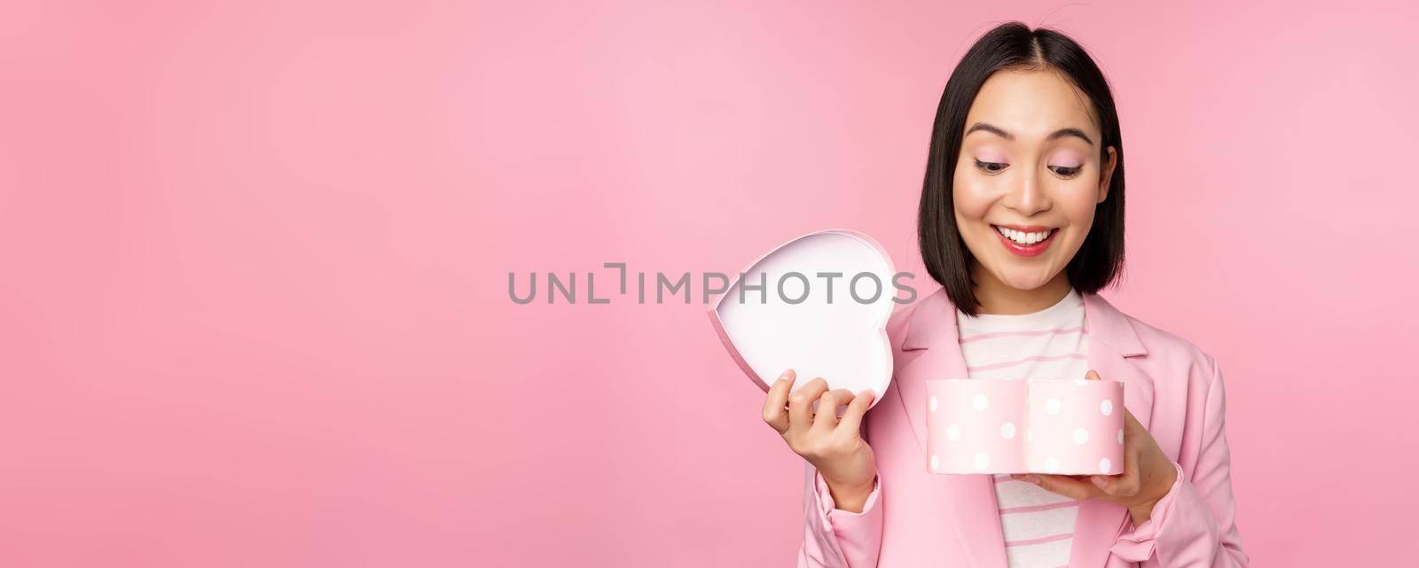 Happy cute korean girl in suit, opens up heart shaped box with romantic gift on white day holiday, standing in suit over pink background.