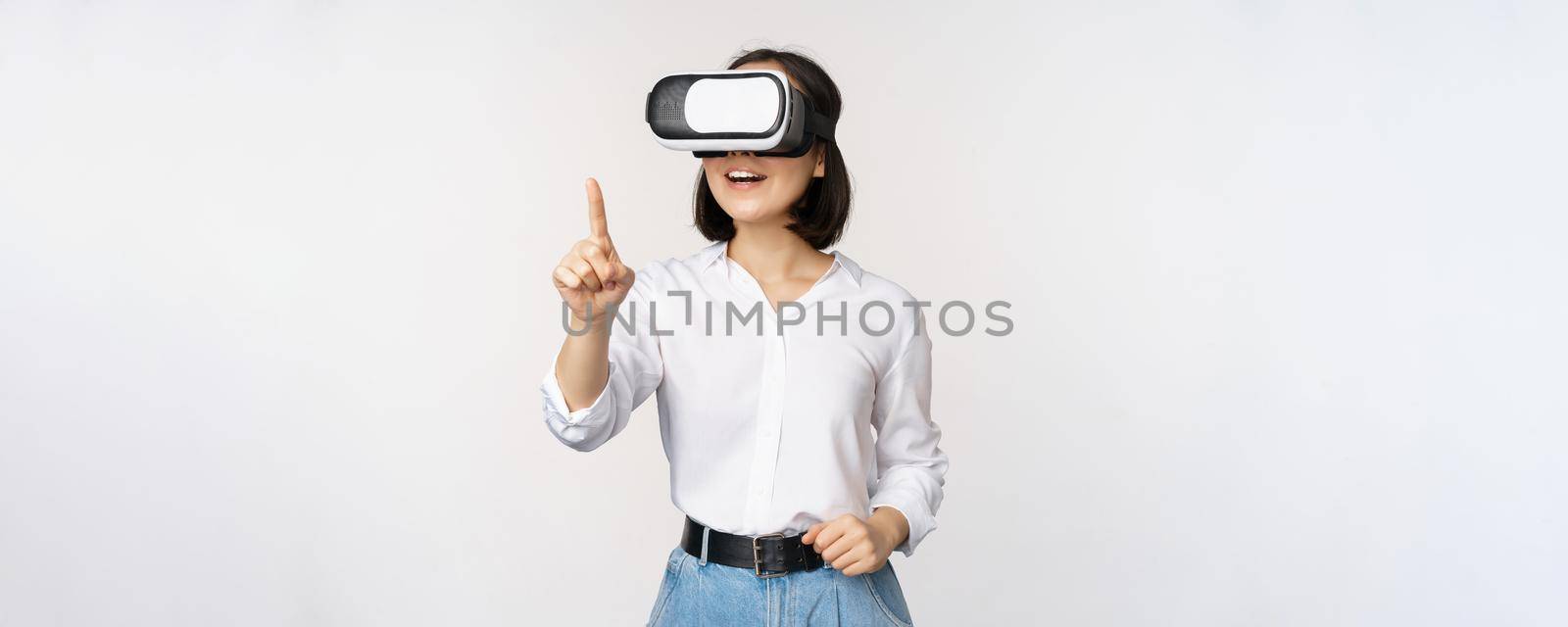 Amazed young asian woman using vr headset. Korean girl in virtual reality glasses pointing at smth and smiling, white background.
