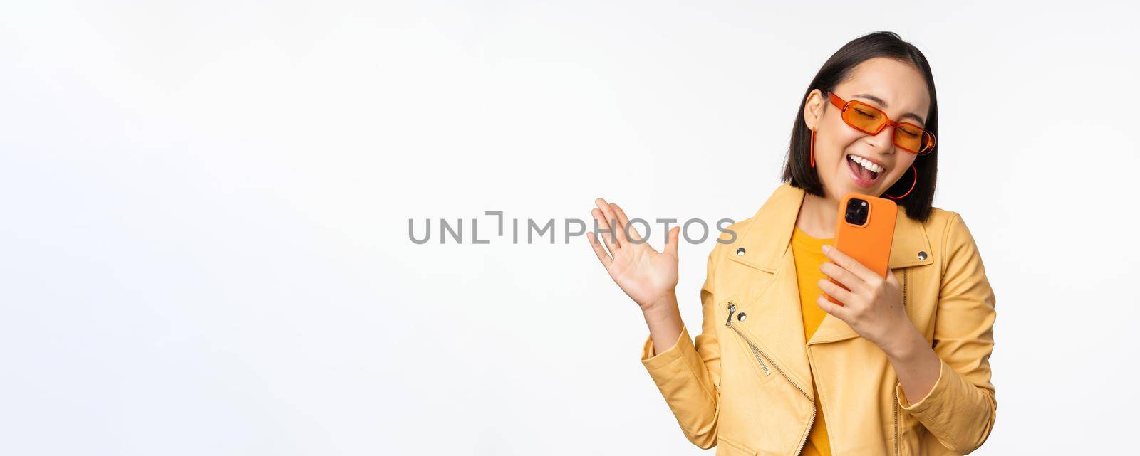 Technology and people concept. Happy dancing asian girl in sunglasses, using smartphone, singing and smiling, standing over white background.