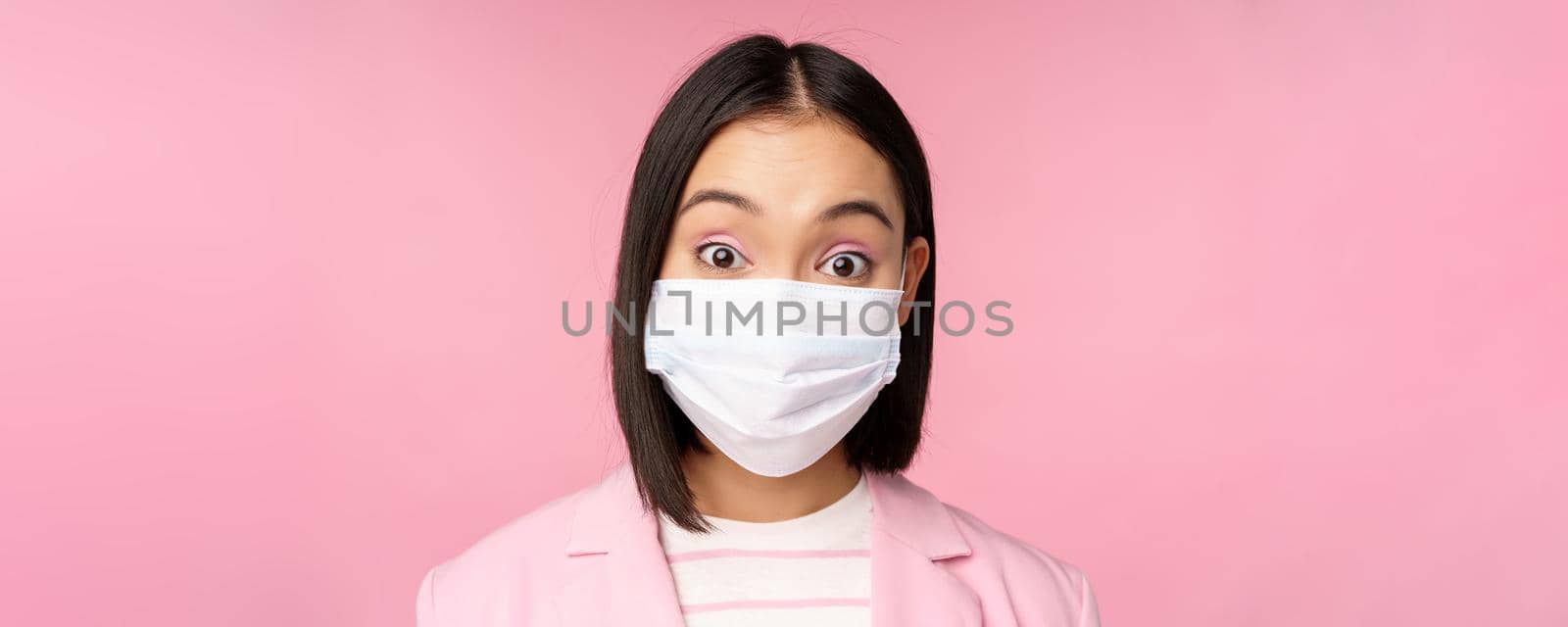 Close-up portrait of asian businesswoman in medical face mask, looking surprised, standing in suit over pink background by Benzoix