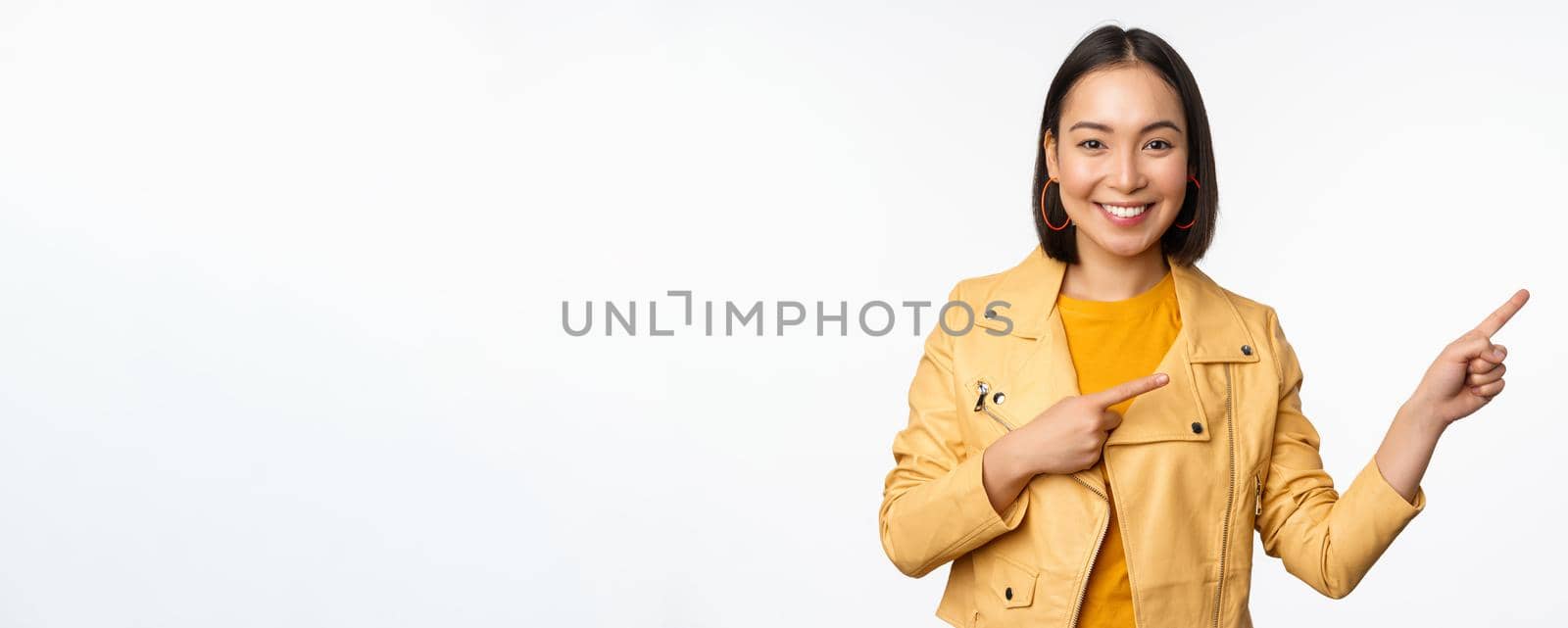 Happy asian woman smiling, pointing fingers right, inviting to check out sale, showing advertisement banner or logo, standing over white background.