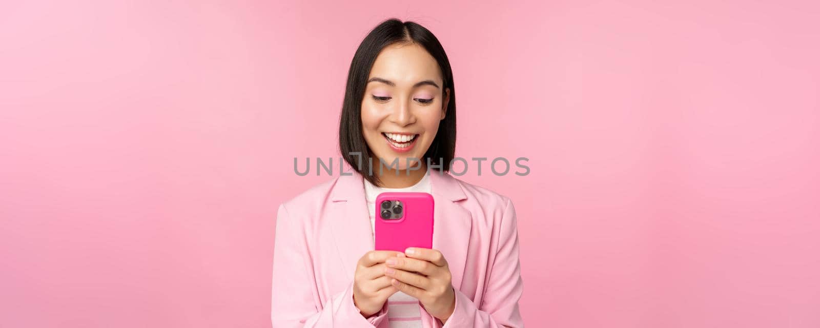 Portrait of asian girl in suit with smartphone, smiling and looking happy, standing over pink studio background by Benzoix