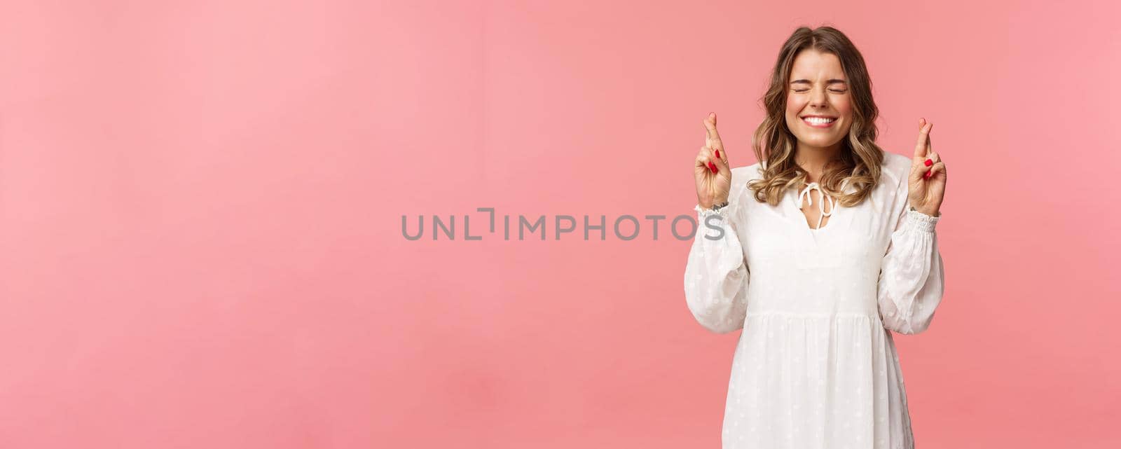 Portrait of hopeful lucky cute blond girl in white dress praying, anticiapte miracle, making wish to win, cross fingers good luck, smiling and close eyes and pleading god, pink background by Benzoix