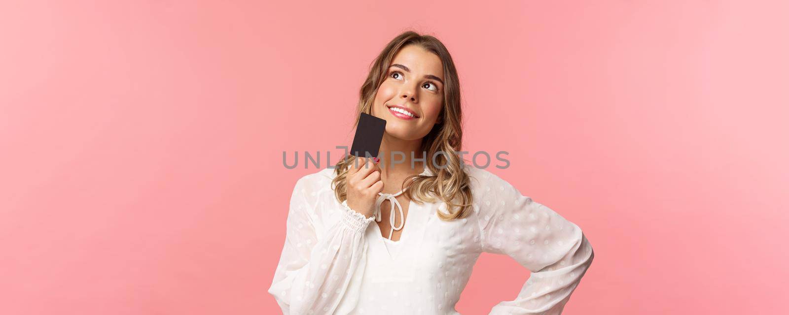 Close-up portrait of thoughtful and creative blond woman in white dress, touching chin with credit card and smiling dreamy as looking up and thinking, picturing what buy as present, pink background.