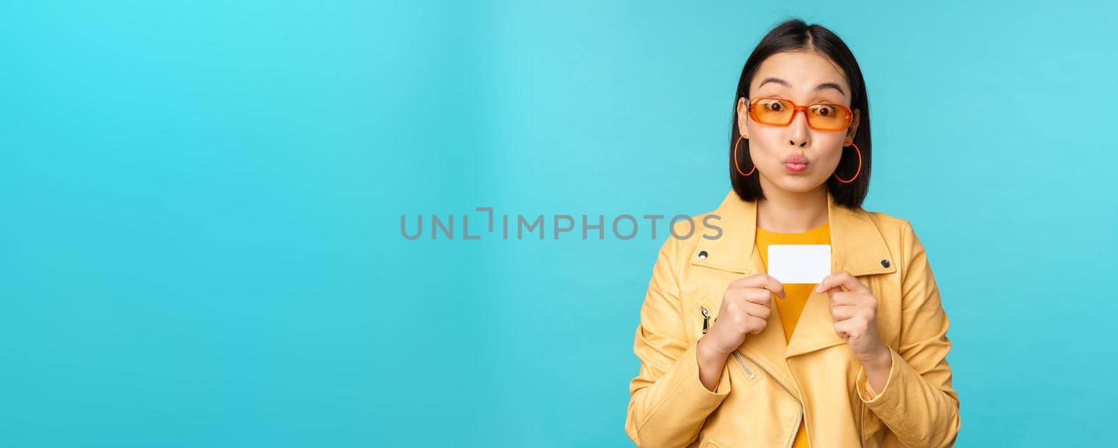Stylish young asian woman in sunglasses, showing credit card and smiling, recommending bank, contactless payment or discounts in store, standing over blue background by Benzoix