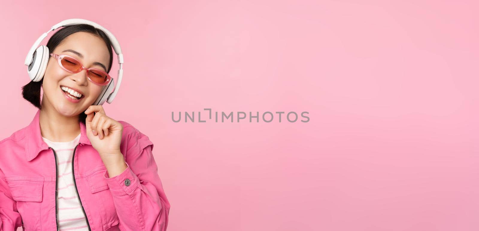 Dancing stylish asian girl listening music in headphones, posing against pink background.