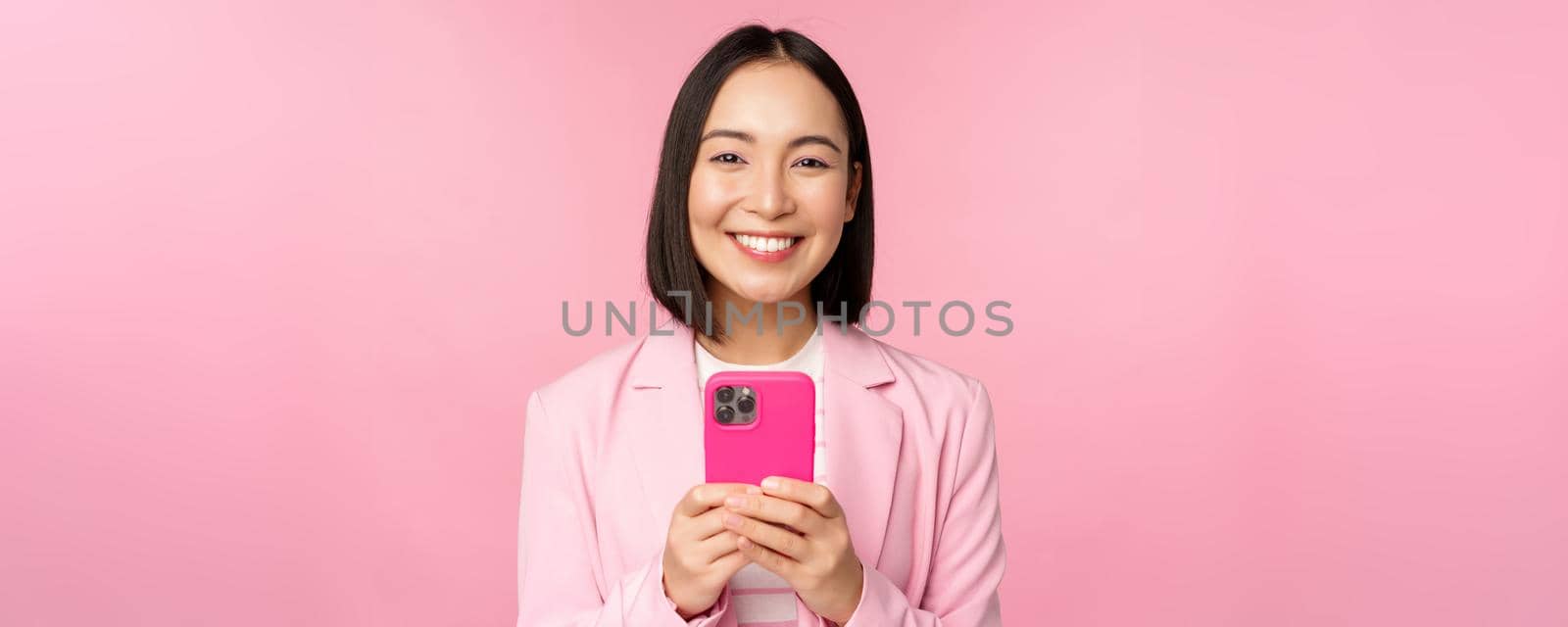Portrait of smiling business woman, asian corporate person using smartphone, mobile phone application, standing over pink background.