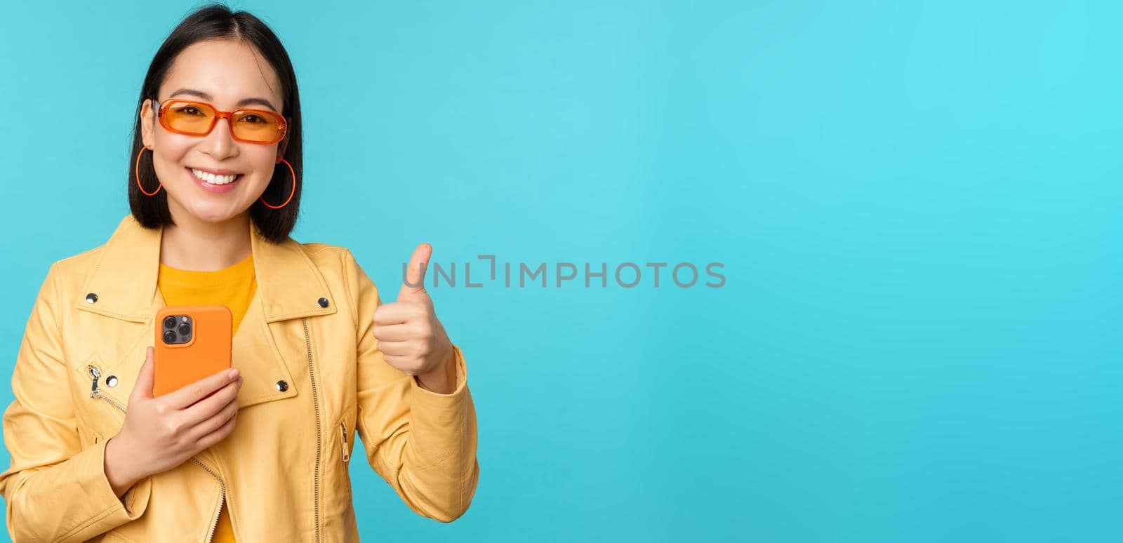 Smiling asian woman showing thumbs up, recording on mobile phone, using smartphone app and recommending it, standing over blue background.