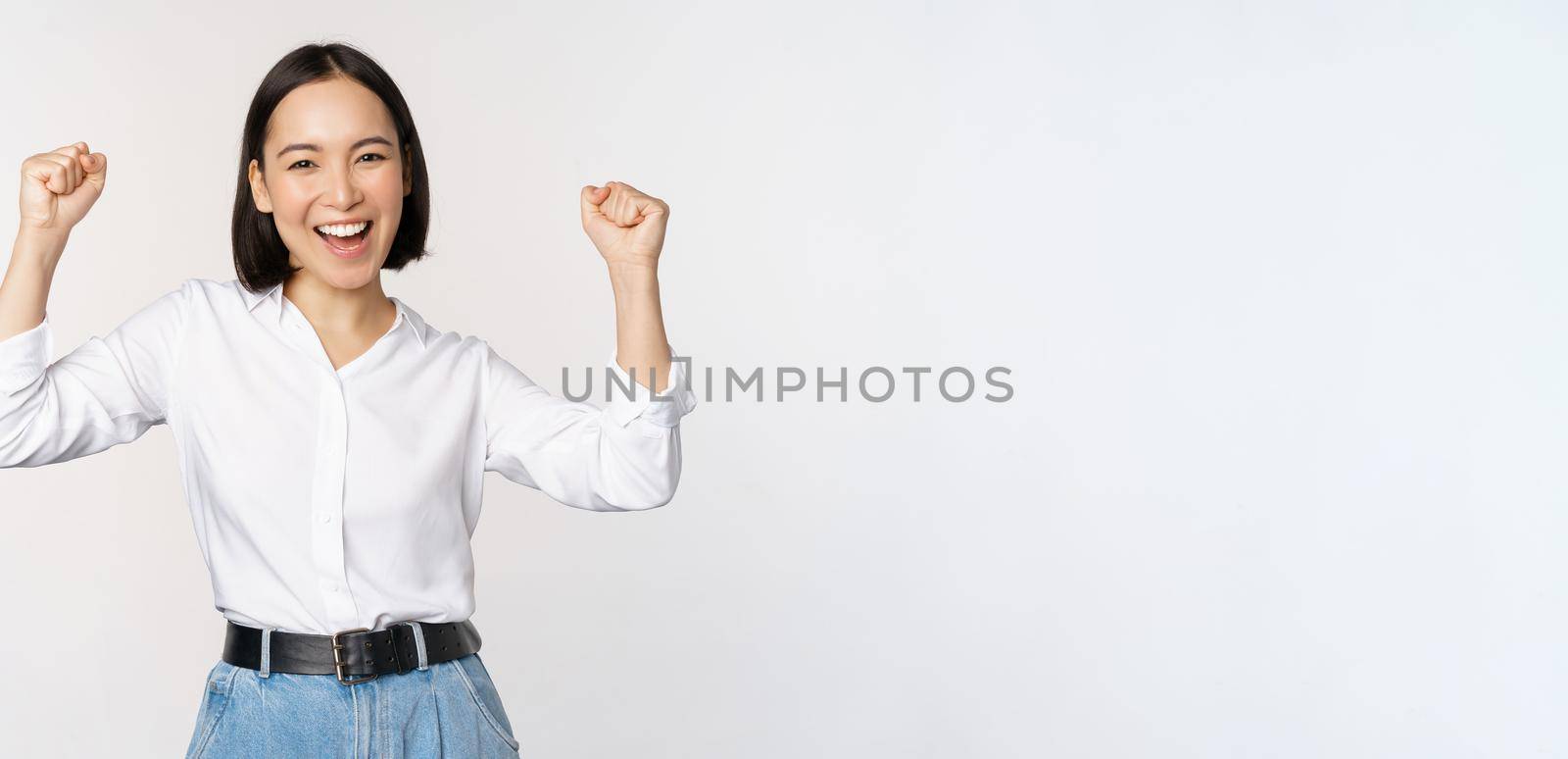 Enthusiastic asian woman rejoicing, say yes, looking happy and celebrating victory, champion dance, fist pump gesture, standing over white background.