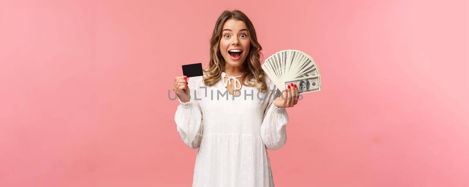 Portrait of excited happy good-looking blond girl in white dress, winning money, placed good bet, made deal, holding dollars money and credit card, smiling amused at camera, pink background by Benzoix