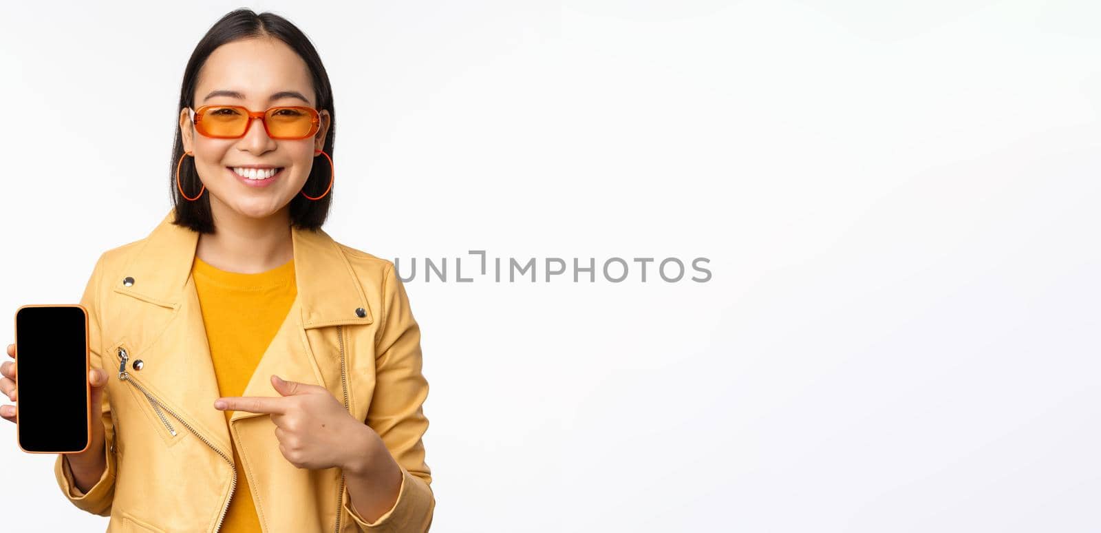 Portrait of stylish korean girl in sunglasses, smiling, pointing finger at smartphone screen, showing mobile phone application, standing over white background.