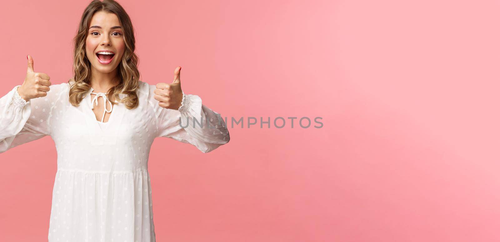 Portrait of feminine blond woman in white dress, show thumbs-up in approval, say yes, approve or agree, satisfied and recommend product, standing pink background in white dress.