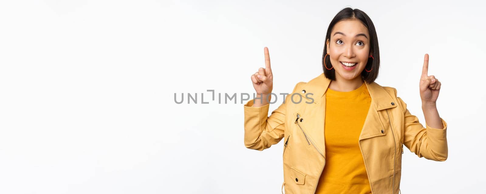 Image of smiling asian brunette woman pointing fingers up, showing advertisement with happy face, posing against white background.