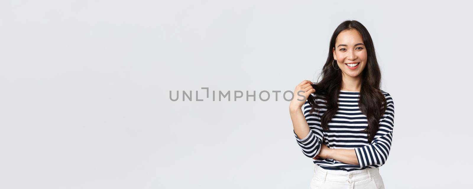 Lifestyle, people emotions and casual concept. Charming smiling korean girl in striped shirt, touching hair strand and grinning happily camera, talking to friend, white background.