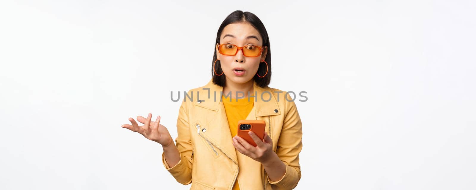 Portrait of korean girl in sunglasses, holding smartphone, looking confused and shrugging, standing over white background. Copy space