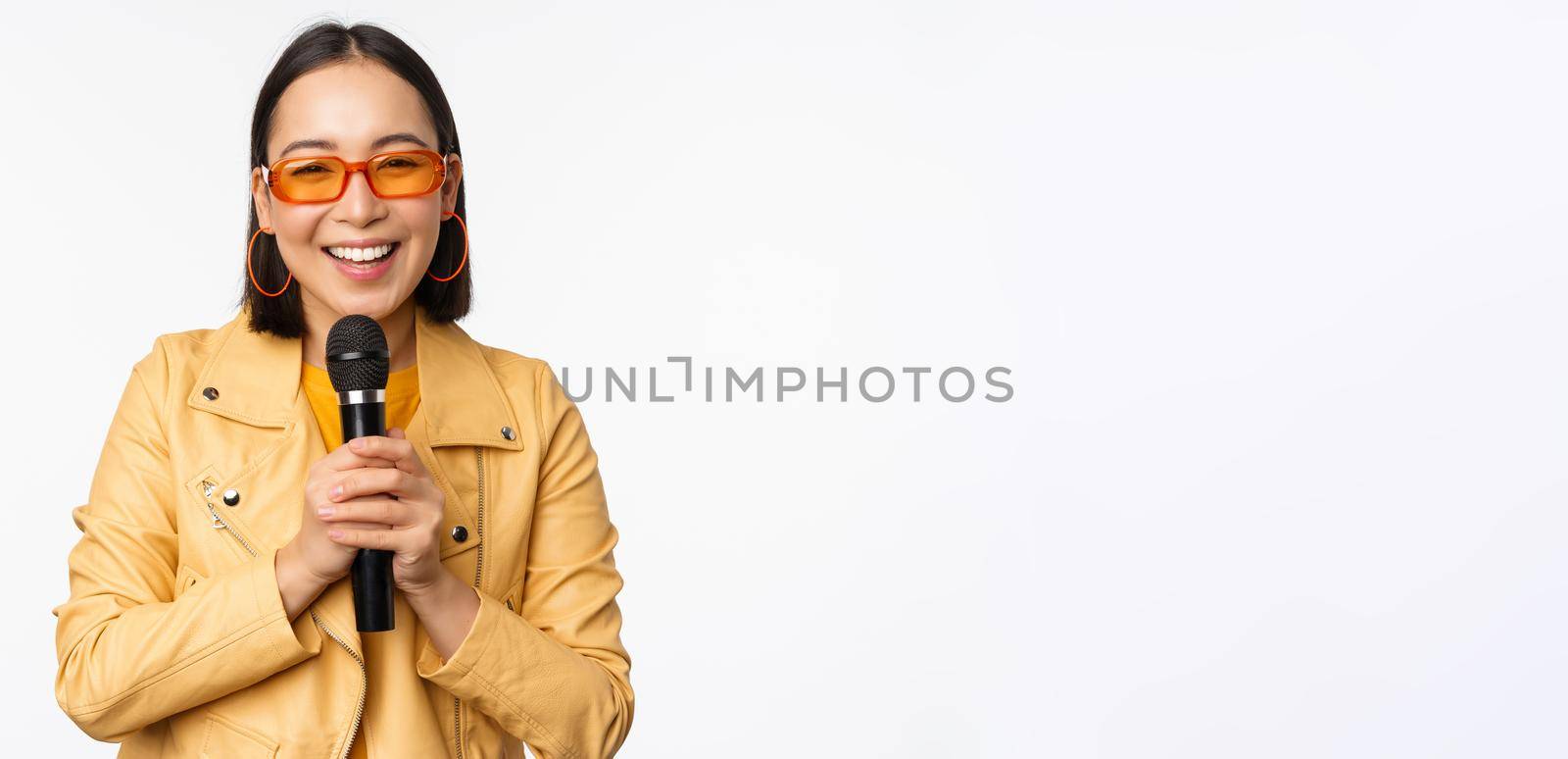 Stylish asian girl in sunglasses, singing songs with microphone, holding mic and dancing at karaoke, posing against white background.