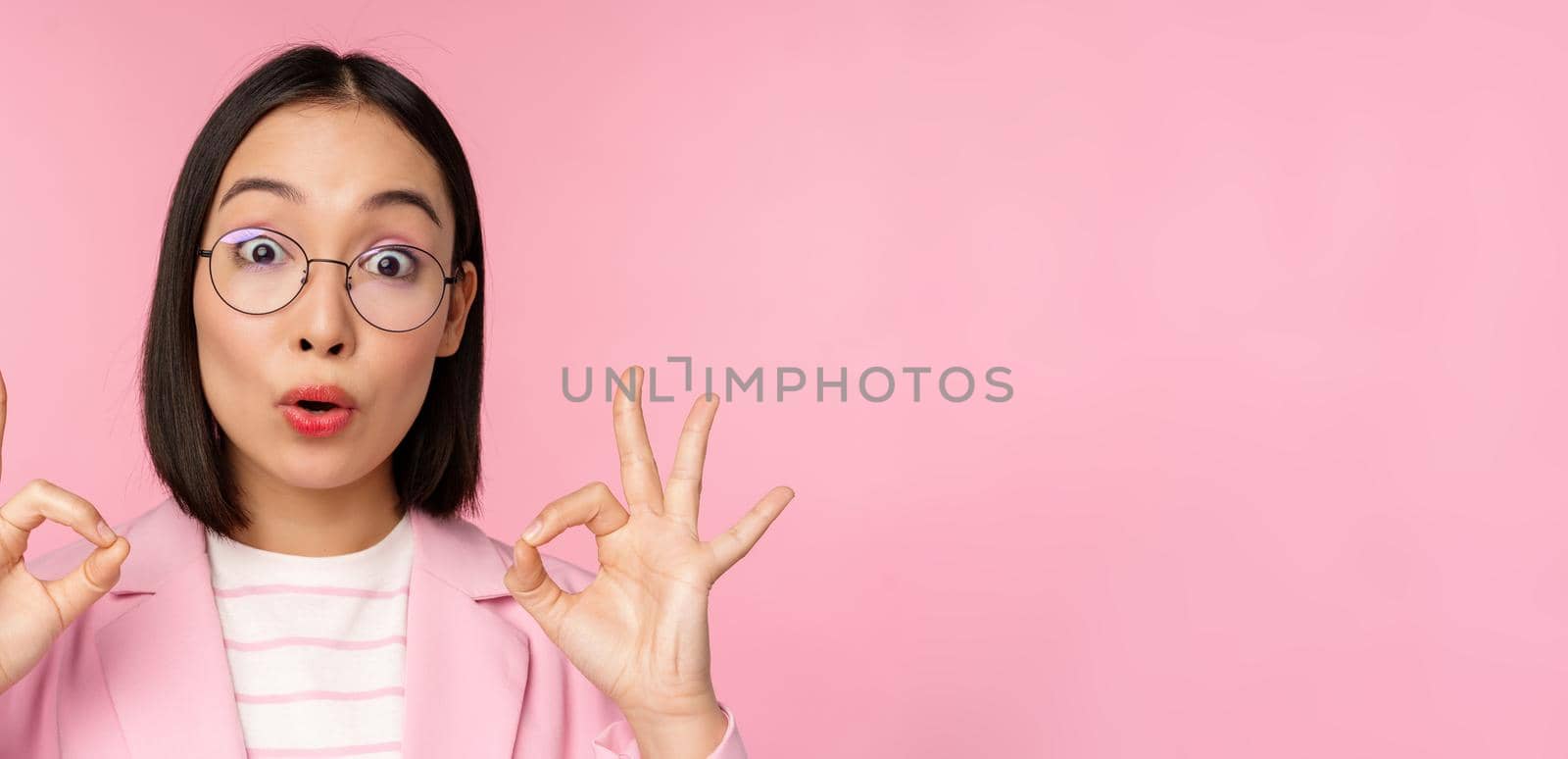 Close up portrait of business woman looks impressed and shows okay sign in approval, recommending company. Young corporate lady in glasses shows ok gesture, pink background.