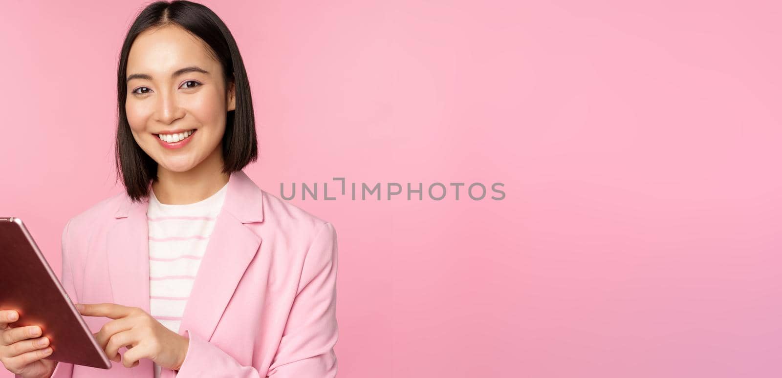 Portrait of young asian corporate woman, office lady with digital tablet, wearing suit, smiling and looking professional, posing against pink background.
