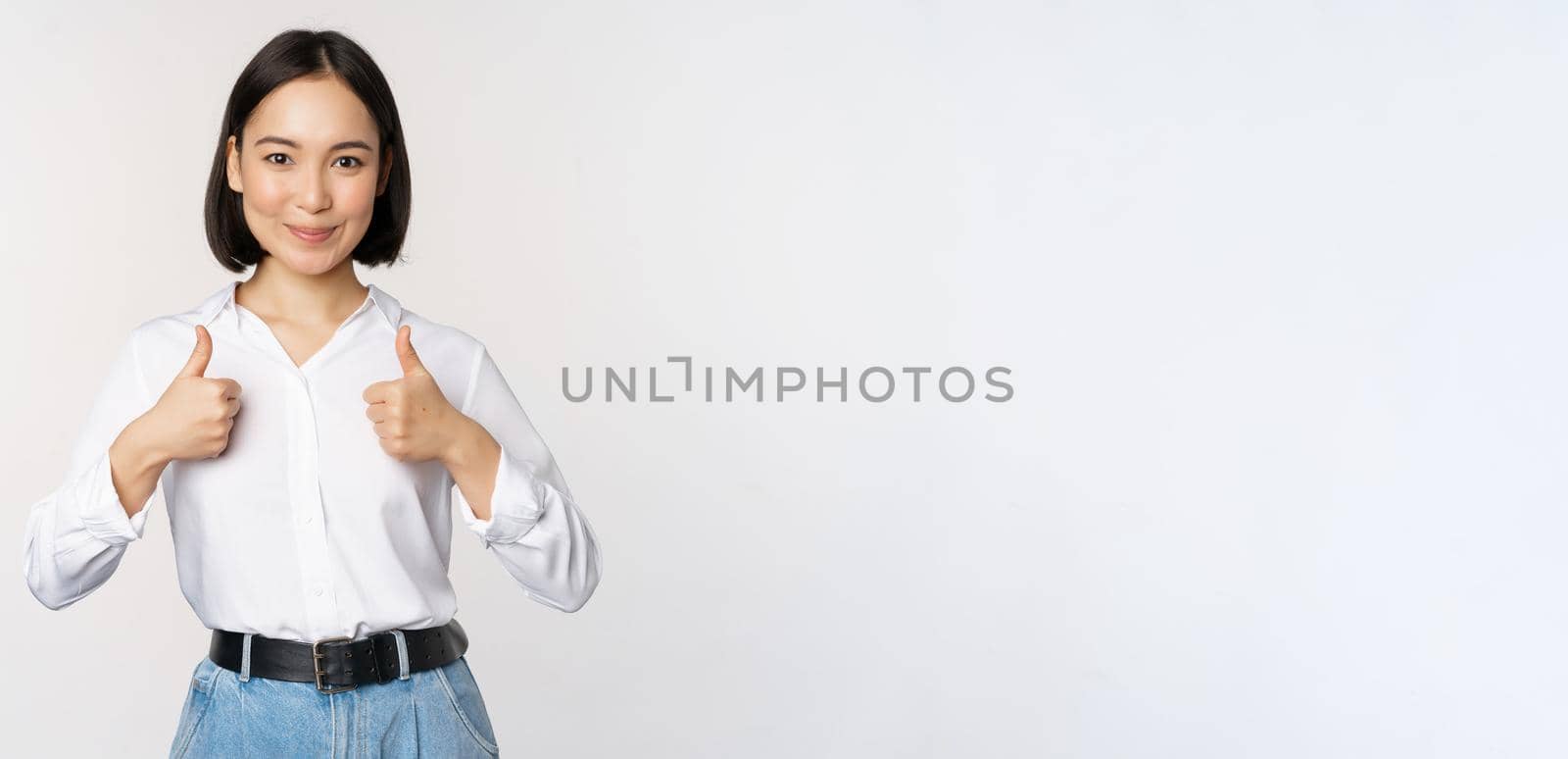 Image of beautiful adult asian woman showing thumbs up, wearing formal office, university clothing, recommending company, standing over white background.