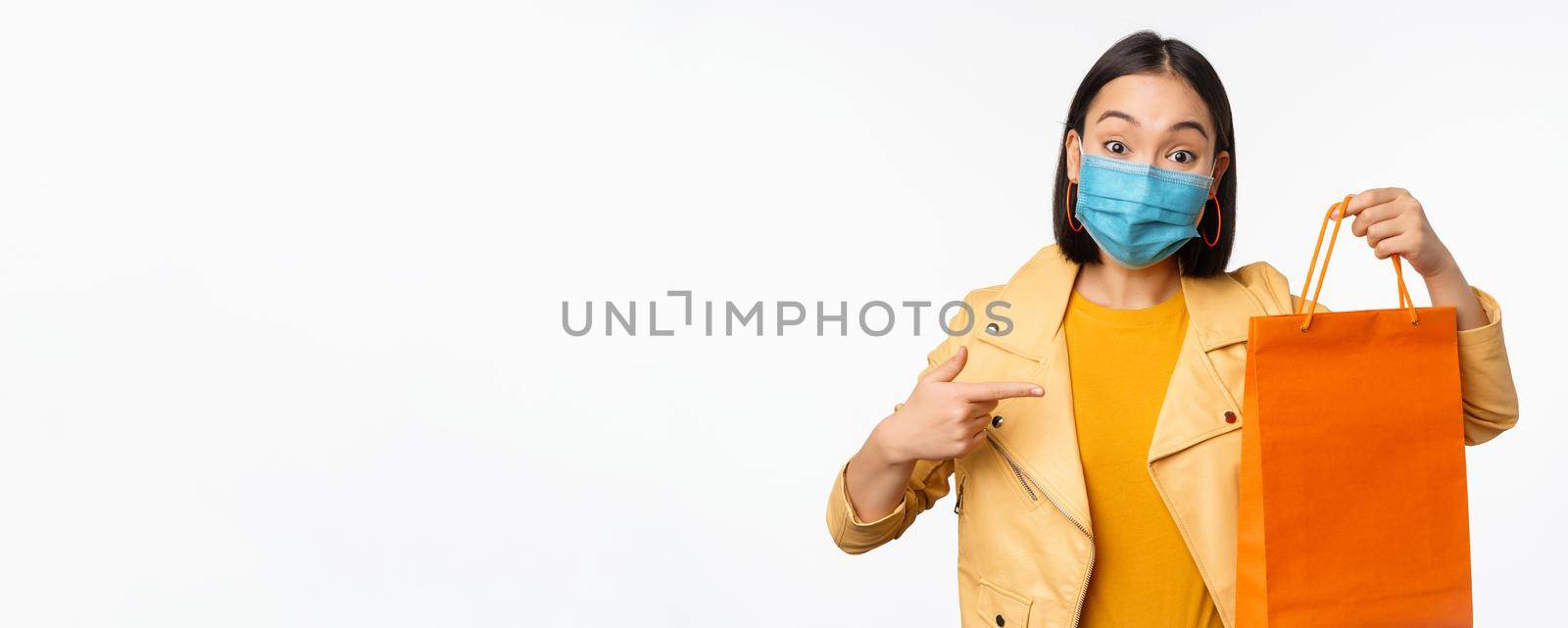 Covid-19 and shop concept. Young asian stylish woman, wearing medical face mask, holding shopping bag, going in malls during pandemic, white background.