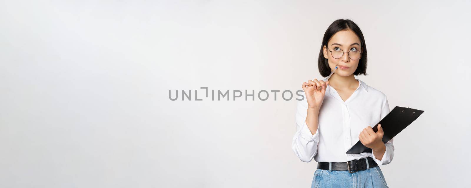 Asian girl in glasses thinks, holds pen and clipboard, writing down, making notes, standing over white background.