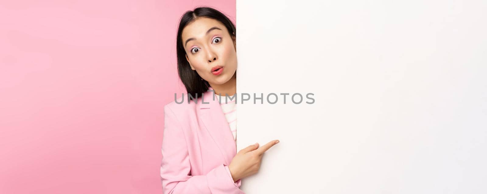 Portrait of young japanese business woman, corporate lady in suit pointing on wall with chart, showing diagram or advertisement on empty copy space, pink background by Benzoix