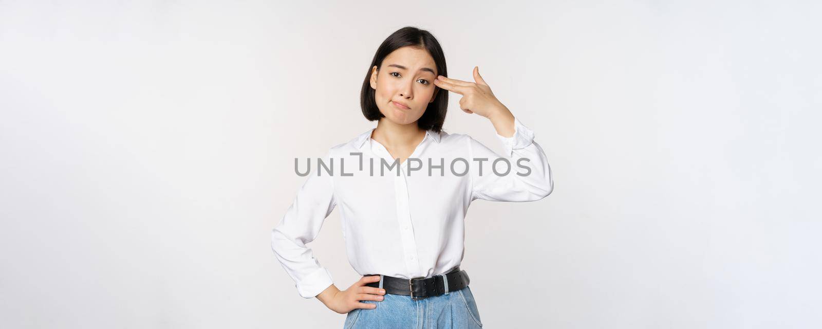 Image of tired asian woman, annoyed by smth, pointing fingers at her head, standing over white background. Copy space