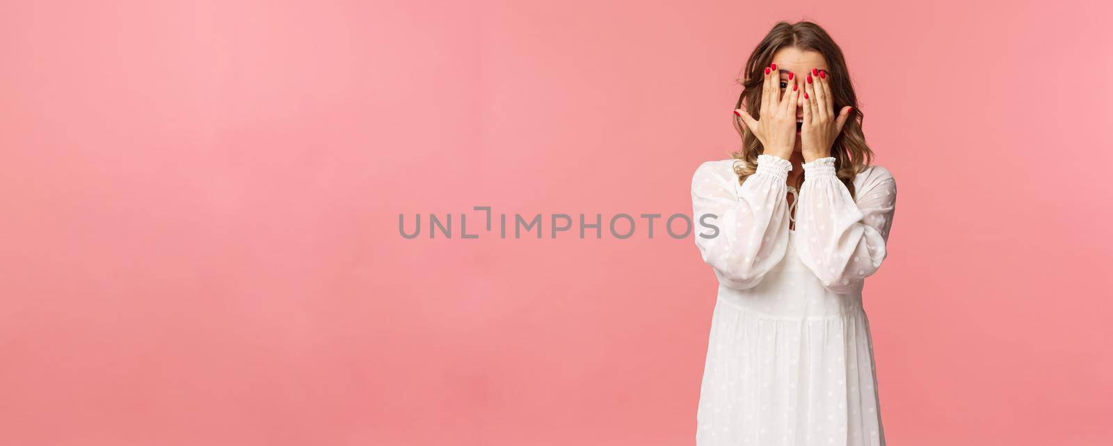Portrait of silly cute girl with blond short hair, wear white dress, hiding face behind hands, promise to wait for signal but peeking through fingers, cant resist temptation, stand pink background.