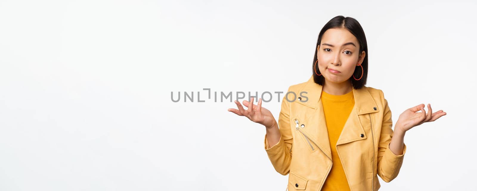 Image of confused young asian woman shrugging shoulders, looking puzzled and clueless at camera, cant understand, standing against white background.