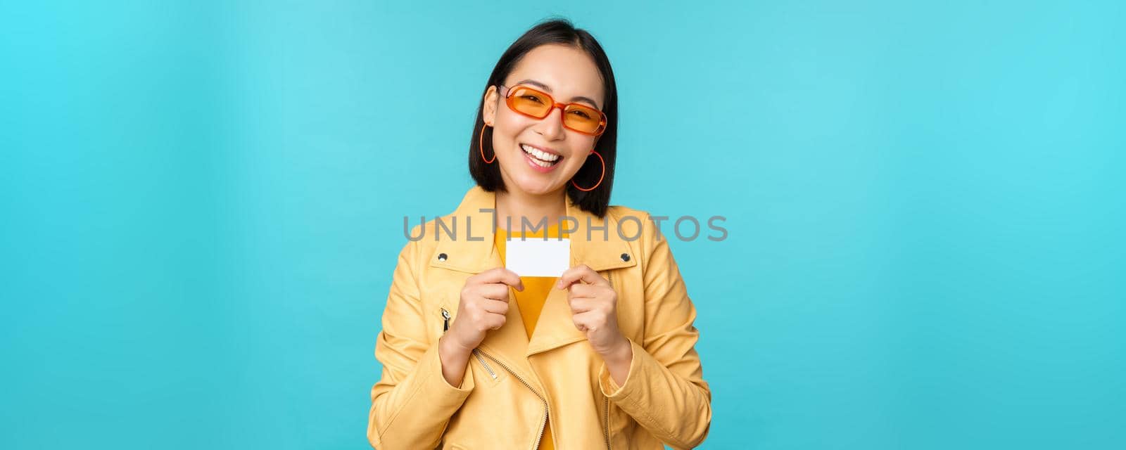 Stylish young asian woman in sunglasses, showing credit card and smiling, recommending bank, contactless payment or discounts in store, standing over blue background.