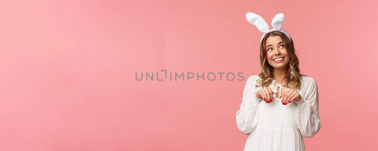 Holidays, spring and party concept. Cute romantic young blond girl imitating bunny, wear white dress lovely rabbit ears, make hand-paws near chest and look dreamy up with daydreaming smile.