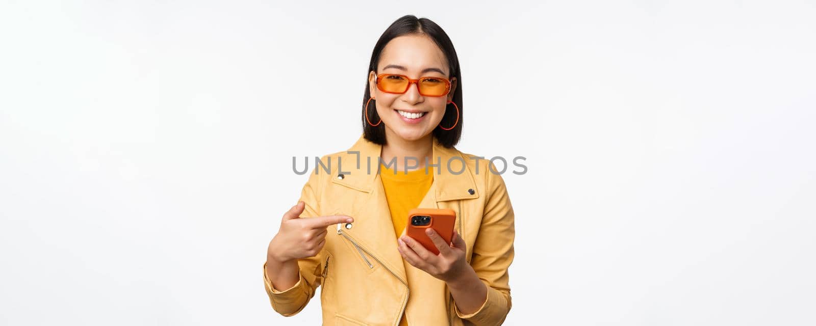 Beautiful smiling asian girl in sunglasses, pointing finger at smartphone, showing app, store on mobile phone, standing over white background.