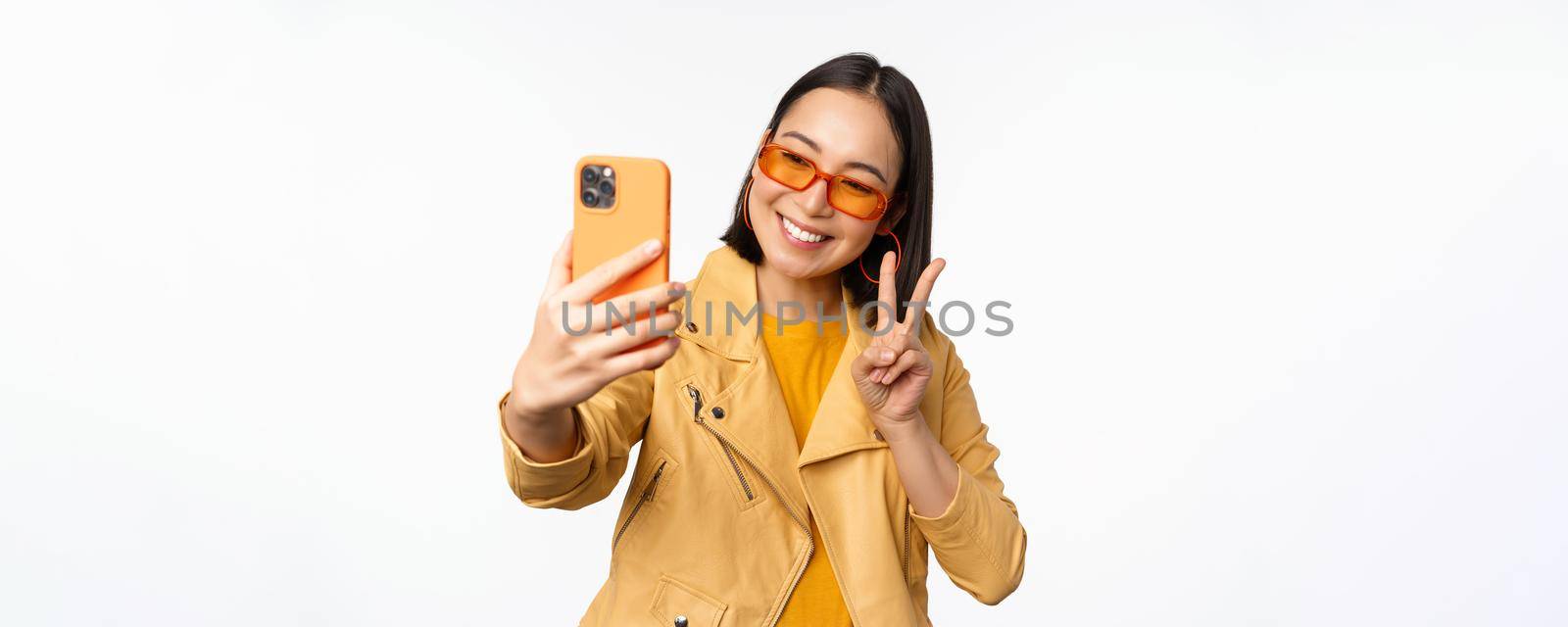 Stylish, beautiful asian girl in sunglasses, taking selfie on smartphone, posing for photo, holding mobile phone, white studio background by Benzoix