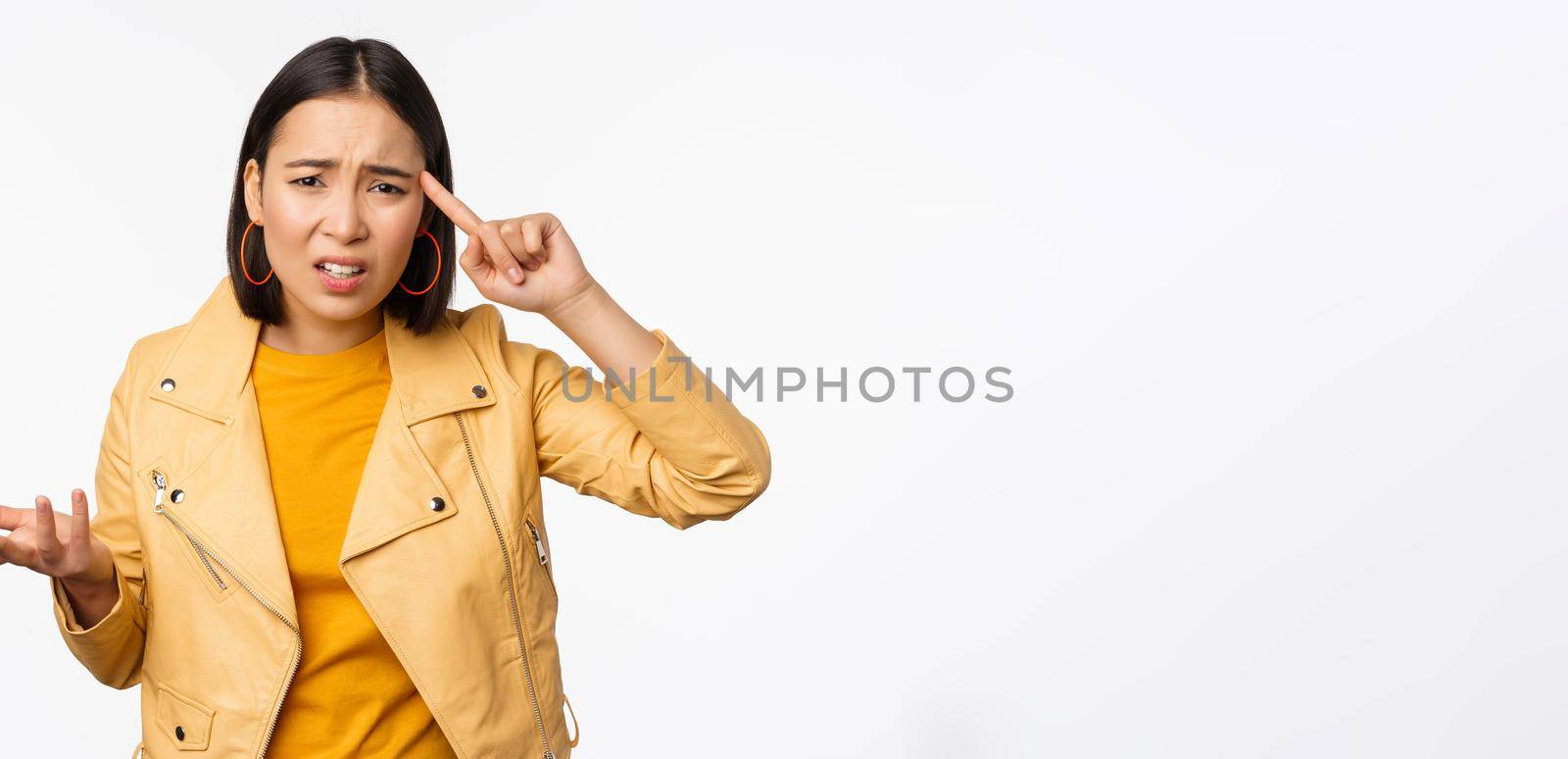 Annoyed asian woman roll finger near temple and staring frustrated, scolding someone stupid or crazy, standing over white background.