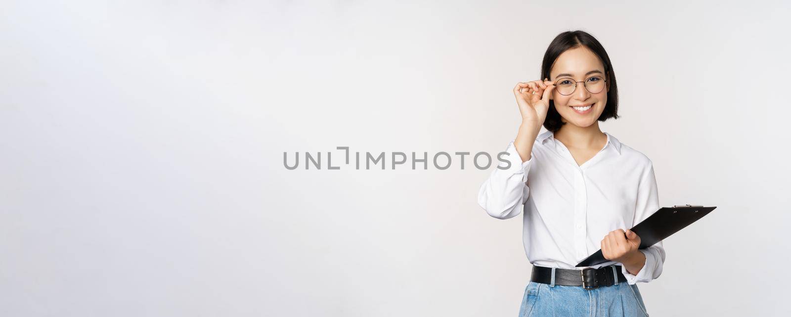 Young woman, office worker manager in glasses, holding clipboard and looking like professional, standing against white background.