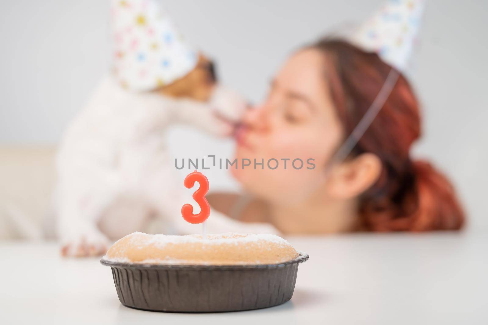 Caucasian woman and jack russell terrier in holiday caps look at the cake with a candle. The dog and the owner celebrate the third birthday. by mrwed54