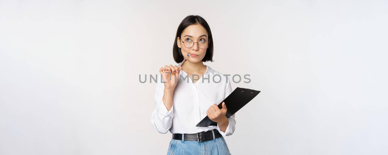 Asian girl in glasses thinks, holds pen and clipboard, writing down, making notes, standing over white background by Benzoix