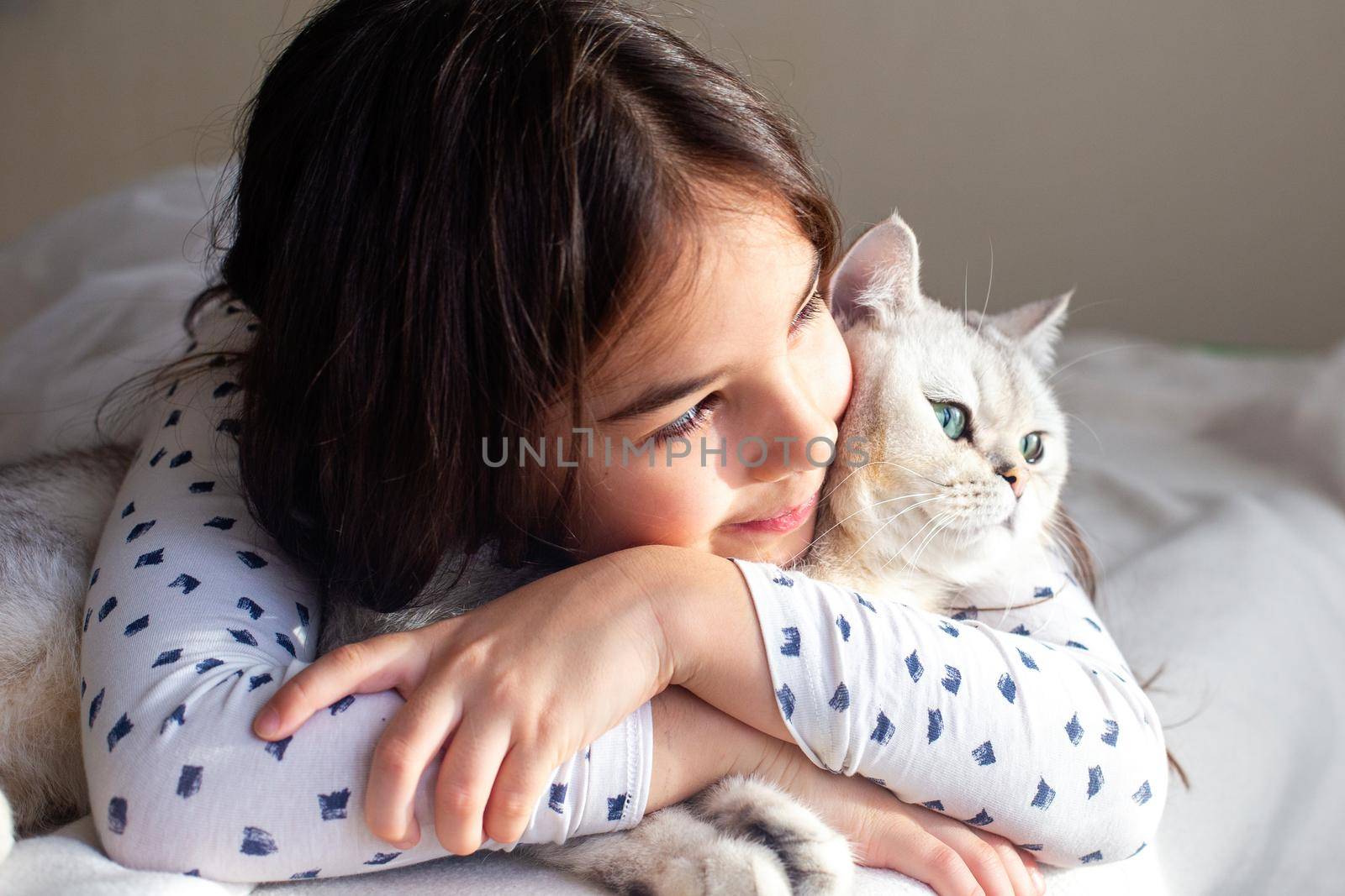 Portrait of little girl lies with a white cat on the bed, hugs him, smiles.