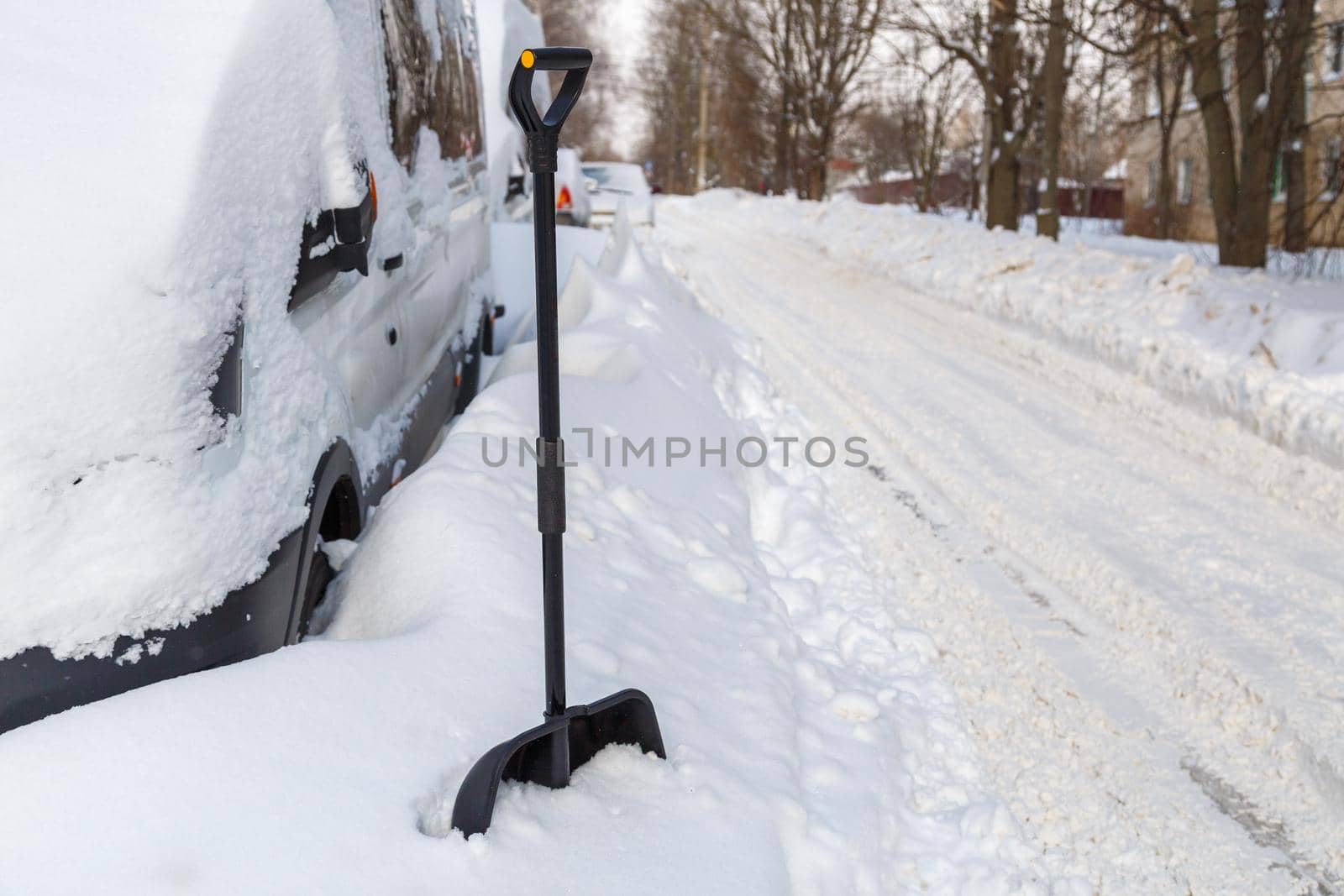 Plastic snow shovel in front of snow-covered minivan at sunny winter morning.