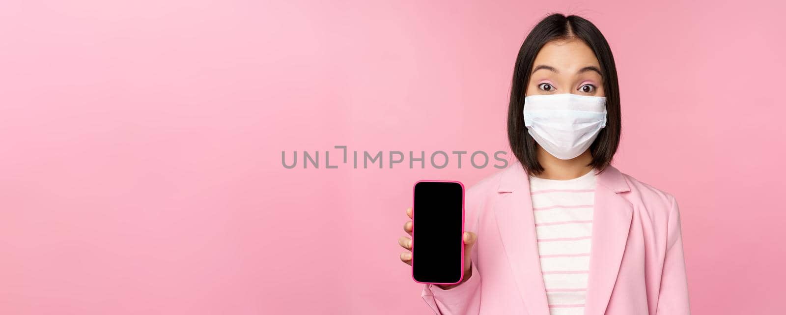 Portrait of smiling korean saleswoman in medical face mask, business suit, showing smartphone screen, standing over pink background.
